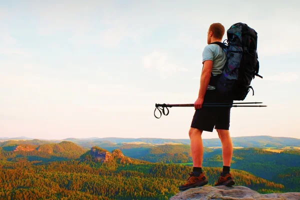 Tall backpacker with poles in hand. Sunny summer evenng in rocky mountains. Hiker with big backpack stand on rocky view point above valley.