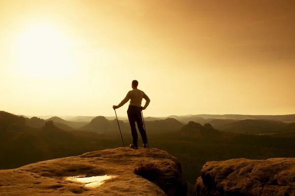 Ginger hair man in grey t-shirt and dark trekking trousers on sharp rock. Tourist with pole above misty valley.