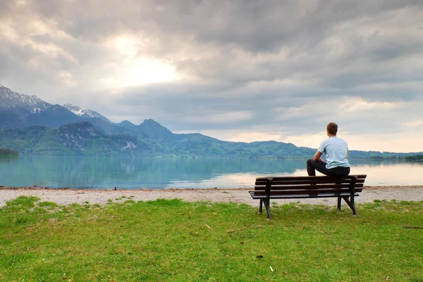 Tired adult man in blue shirt sit on old wooden bench at mountain lake coast