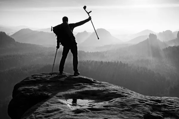 Tourist with  medicine crutch above head achieved mountain peak. Hiker with broken leg in immobilizer.  Deep misty valley bellow silhouette of man with hand in air. Spring daybreak