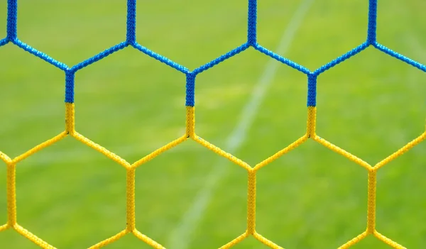 Detail of yellow blue crossed soccer nets, soccer football in goal net with grass on playground in background. Honeycomb shape.