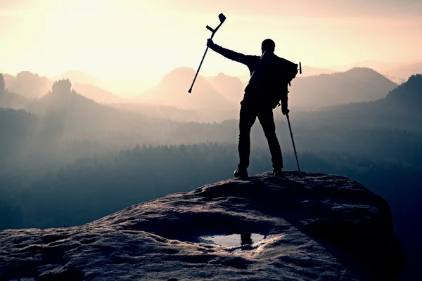 Hiker with crutch above head. Deep valley bellow man silhouette