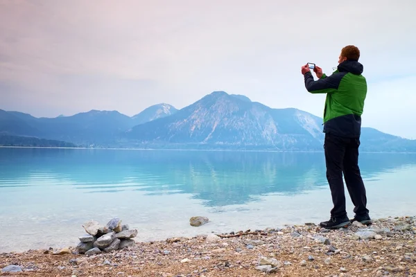 Tall man hold cellphone, take photo of autumn mountain lake scenery