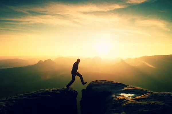 Crazy hiker umping between rocks. Marvelous colorful daybreak in rocky mountains, heavy orange mist in deep valley. Miracle of nature