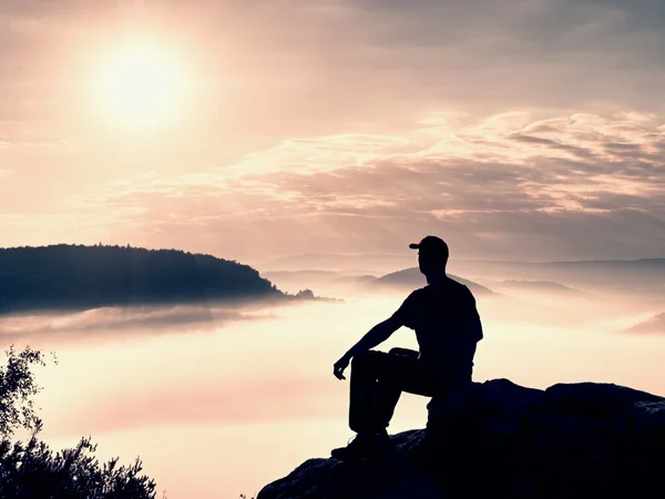 Moment of loneliness. Man sit on rock and watching into colorful mist and fog in valley.