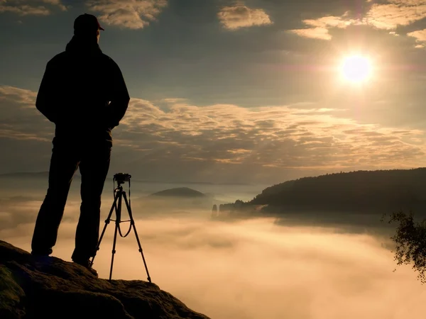 Photographer  on cliff. Nature photographer takes photos with mirror camera on peak of rock. Dreamy fogy landscape, spring orange pink misty sunrise in a beautiful valley below.