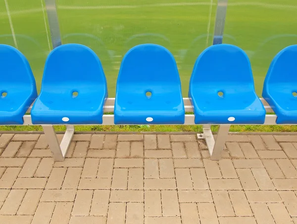 New blue plastic seats on outdoor stadium players bench, chairs with new paint below transparent plastic roof. Plastic football green turf playground.