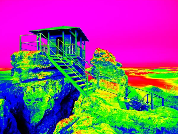Infrared photo, wooden cabin on main peak of rock as view point, dark clouds in the sky. Summer early morning in sandstone rocks. Amazing thermography colors.