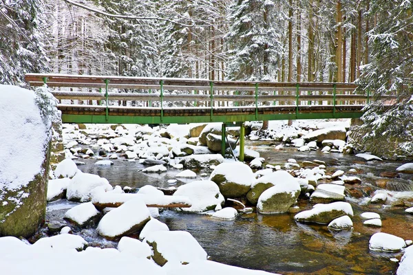 Winter at river and old footbridge. Big stones in stream covered with fresh powder snow and lazy water with low level. Reflections of forest  in water level.