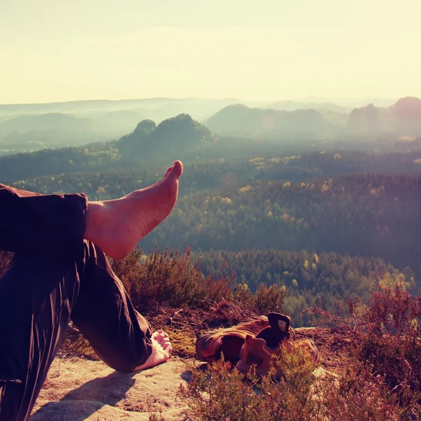 Naked male hairy legs in dark hiking trousers take a rest on peak of rock above valley.