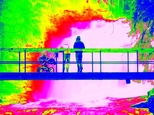 Foamy water of waterfall, bellow footpath bridge with people. Cold water of mountain river in infrared photo. Amazing thermography.