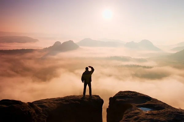 Beautiful moment the miracle of nature. Man stands on the peak of sandstone rock in national park Saxony Switzerland and watching over the misty and foggy morning valley to Sun.