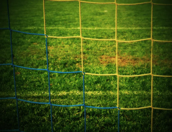 Detail of yellow blue crossed soccer nets, soccer football in goal net with green grass on playground in the background.