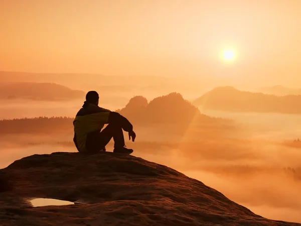 Spring landscape. Hiker in black on the rocky peak. Wonderful daybreak in mountains, heavy orange mist in deep valley. Man sit on the rock and watch over the fog to miracle of nature