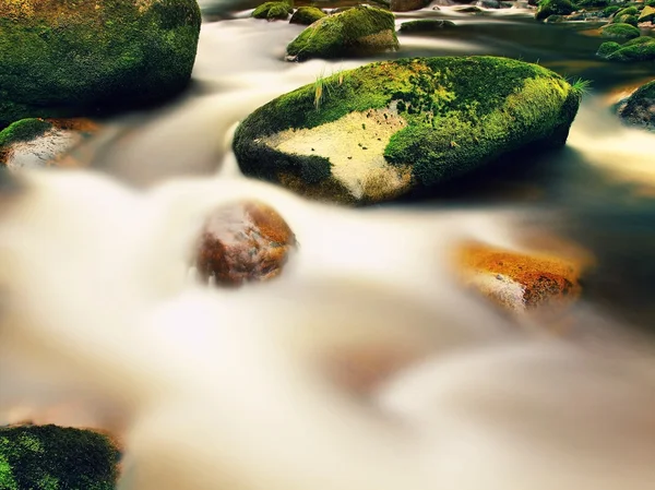 Mountain river with blurred waves of clear water. White curves in rapids between mossy boulders and bubbles create trails.