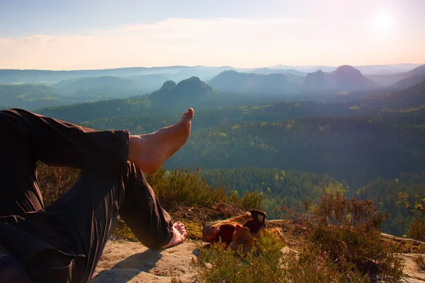 Naked male hairy legs in dark hiking trousers take a rest on peak of rock above valley.