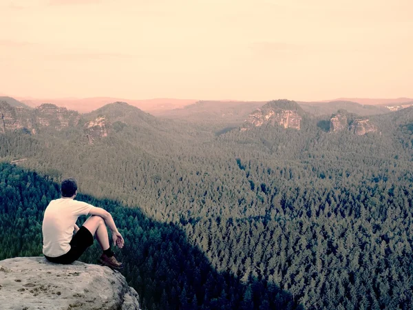 Moment of loneliness. Man in shirt sit on the cliff of rock and watching into forest valley.