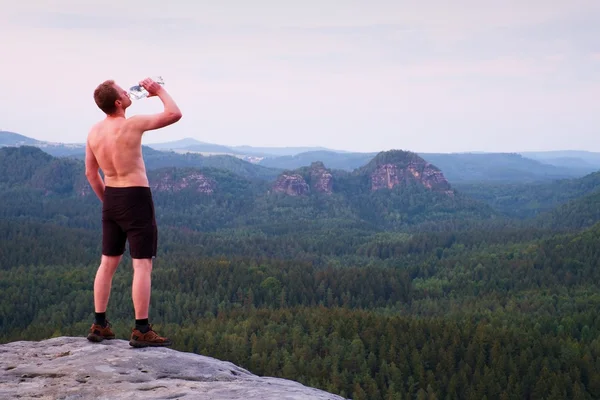 Thirsty hiker in black pants with bottle of water. Sweaty tired tourist on the peak of sandstone rocky park Saxony Switzerland watching into misty landscape.