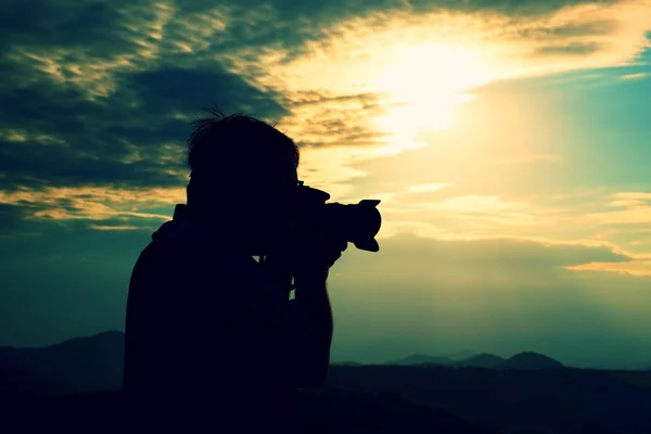Professional photographer in jeans and shirt  takes photos with mirror camera on peak of rock. Dreamy landscape, orange  Sun at horizon