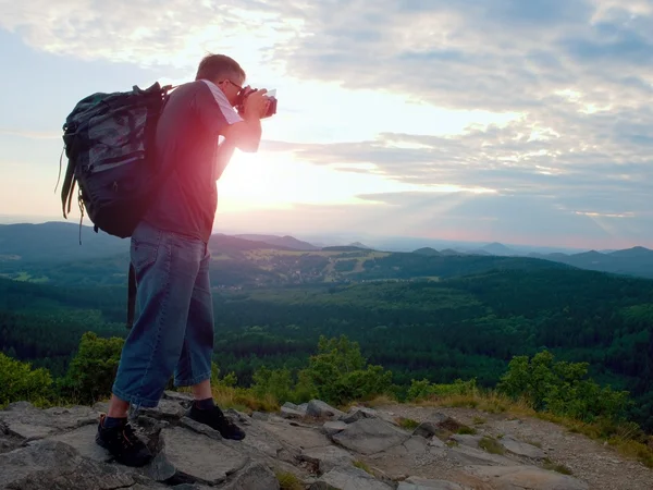 Photographer with big mirror camera on neck and backpack stay on peak of rock. Hilly landscape, fresh green color in valley.