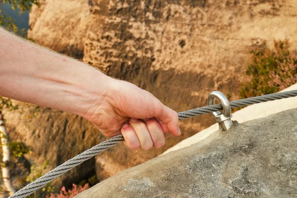 Rock climber's hands hold on steel twisted rope at steel bolt eye anchored in rock. Tourist path via ferrata.