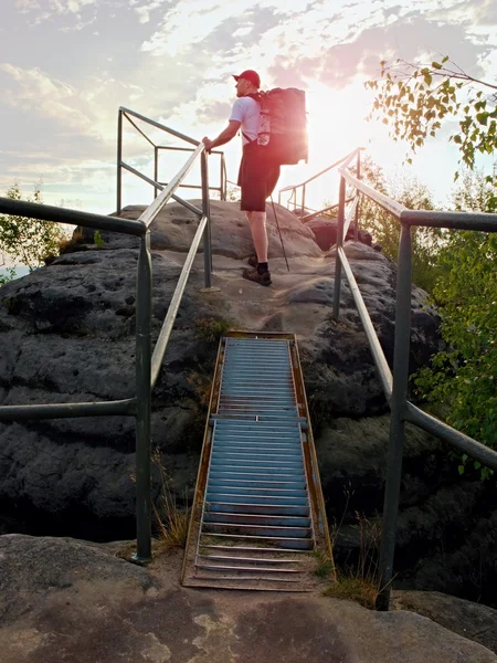 Tall  backpacker hold handrail on rock. Sunny daybreak in rocks. Hiker with big backpack, baseball cap, dark pants and white shirt.