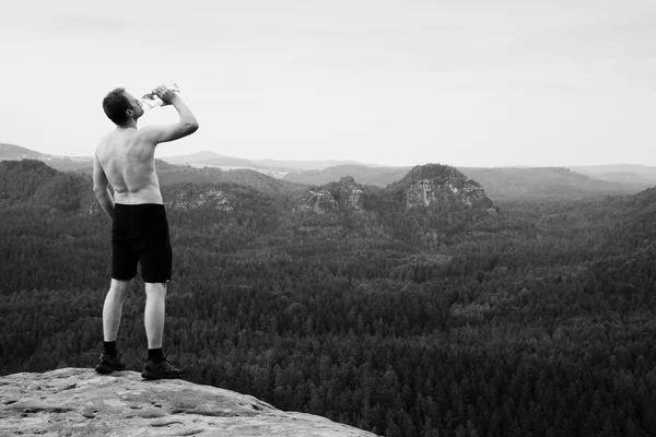 Thirsty hiker in black pants with bottle of water. Sweaty tired tourist on the peak of sandstone rocky park Saxony Switzerland watching into misty landscape.