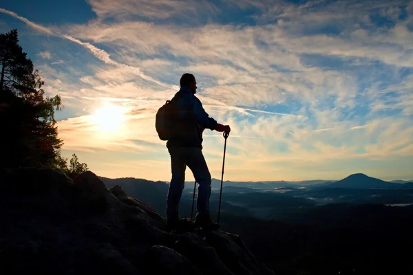 Silhouette of alone hiker with poles in hand. Tourist with sporty backpack stand on rocky view point above misty valley. Sunny spring daybreak in rocky mountains.