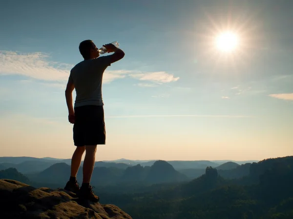 Thirsty hiker in black pants drinks from bottle of water. Sweaty tired tourist on the peak of sandstone rocky park Saxony Switzerland