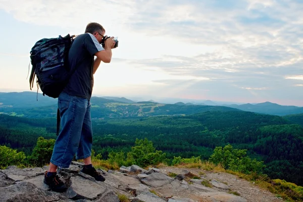 Photographer with big mirror camera on neck and backpack stay on peak of rock. Hilly landscape, fresh green color in valley.
