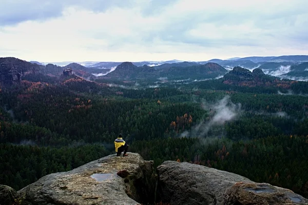 Rear view of tall hiker in yellow black jacket on rocky peak while enjoying a daybreak above fall mountains valley