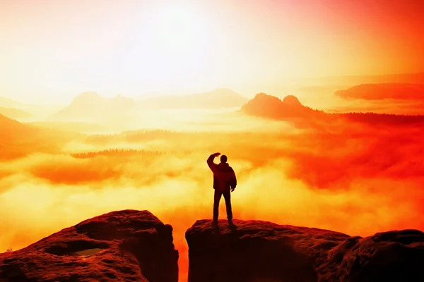 Beautiful moment the miracle of nature. Man stands on the peak of sandstone rock in national park Saxony Switzerland and watching over the misty and foggy morning valley to Sun.