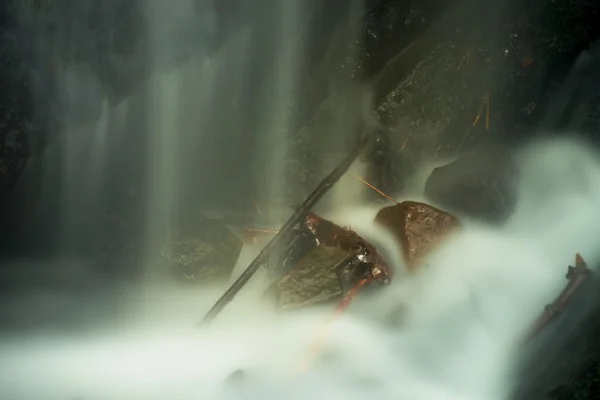 Water spray below small waterfall on mountain stream, water is falling over mossy boulder. The spray create on level and gravel milky water. Broken branches in water