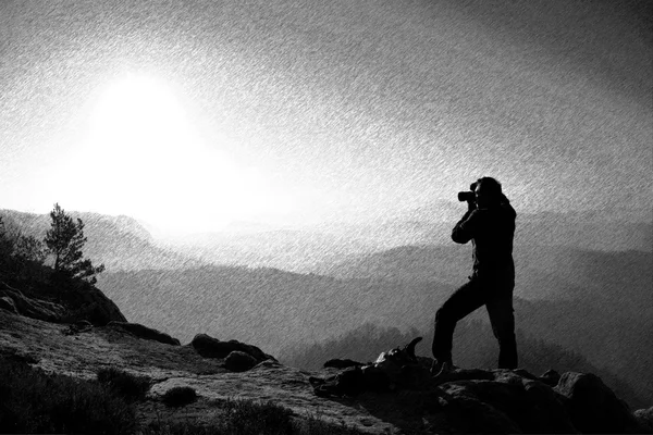 Black and white dashed retro sketch. Professional photographer takes photos with mirror camera on peak of rock.