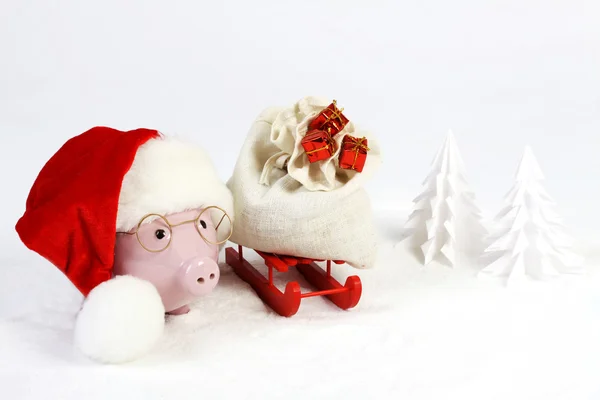 Pink piggy bank with Santas hat with pompom  and glasses standing next to red sled with Santas bag with  three gifts with gold bow and next to snowbound tree on snow