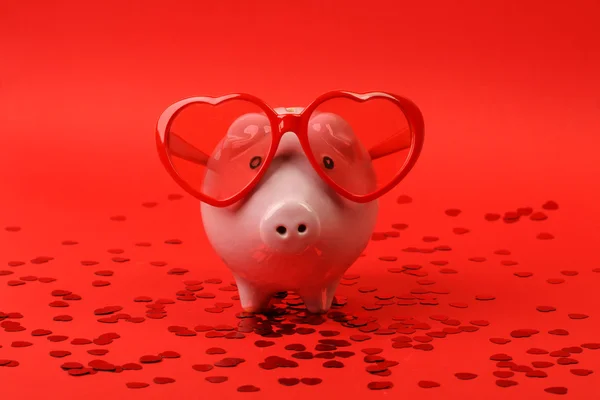 Piggy bank in love with red heart sunglasses standing on red background with shining red heart glitters