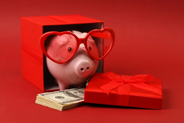 Piggy bank in love with red heart sunglasses standing in gift box with ribbon and with stack of money american hundred dollar bills on red background