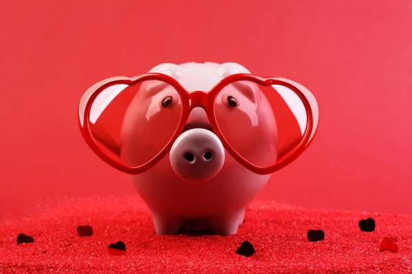 Fallen in love piggy bank with red heart sunglasses standing on red sand with red shining heart glitters in front of red background