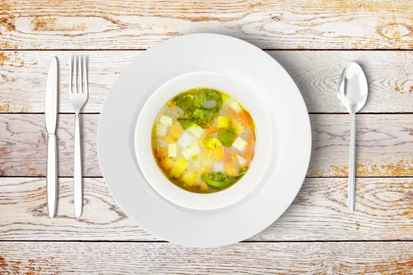 Minestrone soup in white bowl with fork, spoon and knife on wood