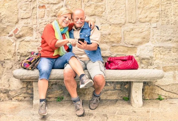 Happy senior couple having fun together with mobile smart phone - Concept of active playful elderly during retirement - Travel lifestyle concept with retired people - Warm cloudy afternoon color tones