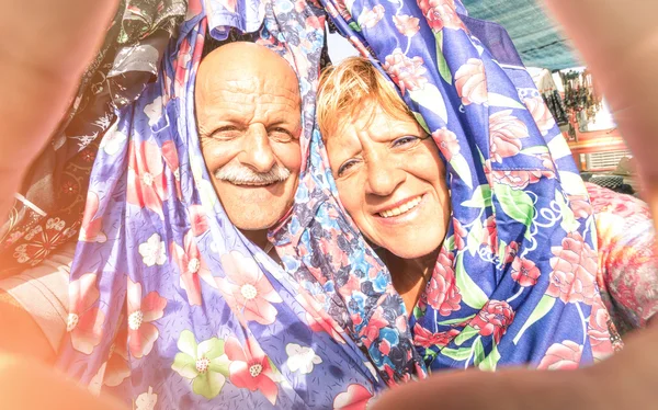 Senior happy couple taking a selfie at the week clothes market traveling around the world - Concept of active elderly and interaction with new technologies and trends