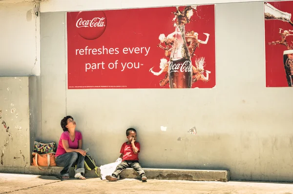 WINDHOEK, NAMIBIA - NOVEMBER 27, 2014: namibian mother and son sitting on floor under a Coca Cola poster. The country has a population of 2.1 million people and a multi-party parliamentary democracy