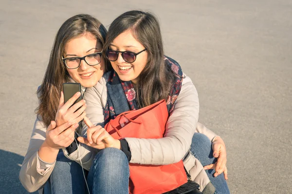 Best friends enjoying time together with smartphone in a spring sunny day - New trends and technology concept with hipster girlfriends having fun outdoors - Alternative four seasons fashion clothes