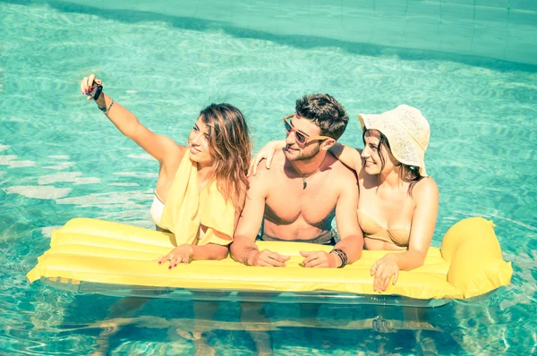 Best friends taking selfie at swimming pool with yellow airbed - Summer friendship concept with new trends and technology - Man and girlfriends with smartphone on a vintage overexposed filtered look