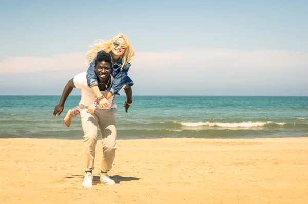 Young multiracial couple at the beach having fun with piggyback jump - Happy mixed race boyfriend and girlfriend playing at the beginning of love story - Multi ethnic integration love against racism