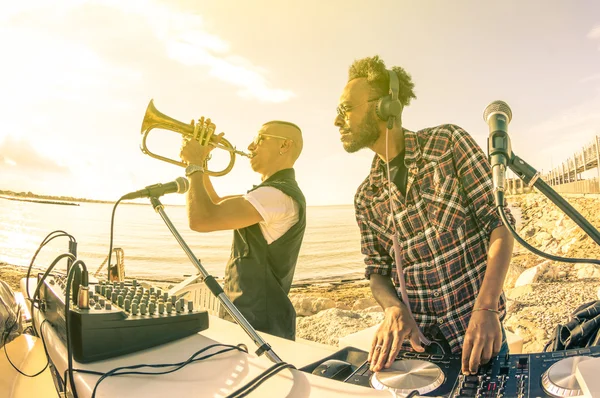 Trendy hipster dj playing summer hits at sunset beach party with trumpet jazz performer - Holidays vacation concept at open air club with house music groove location - Warm vintage sunshine filter
