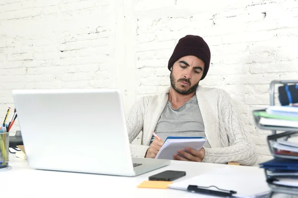 Trendy businessman in cool hipster beanie writing on pad working in at modern home office with computer