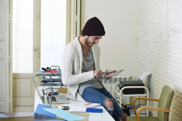 Young trendy businessman in beanie and cool hipster informal look sitting on home office desk using digital tablet pad happy