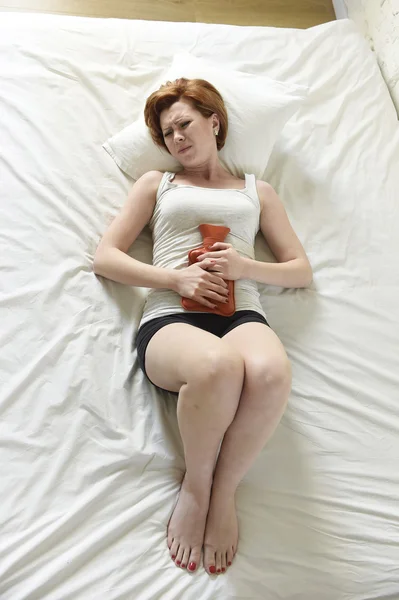 Young woman suffering stomach cramps on belly holding hot water bottle against tummy