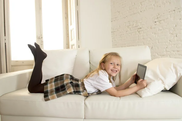 Blond little girl lying on home sofa couch using internet app on digital tablet pad on digital tablet pad